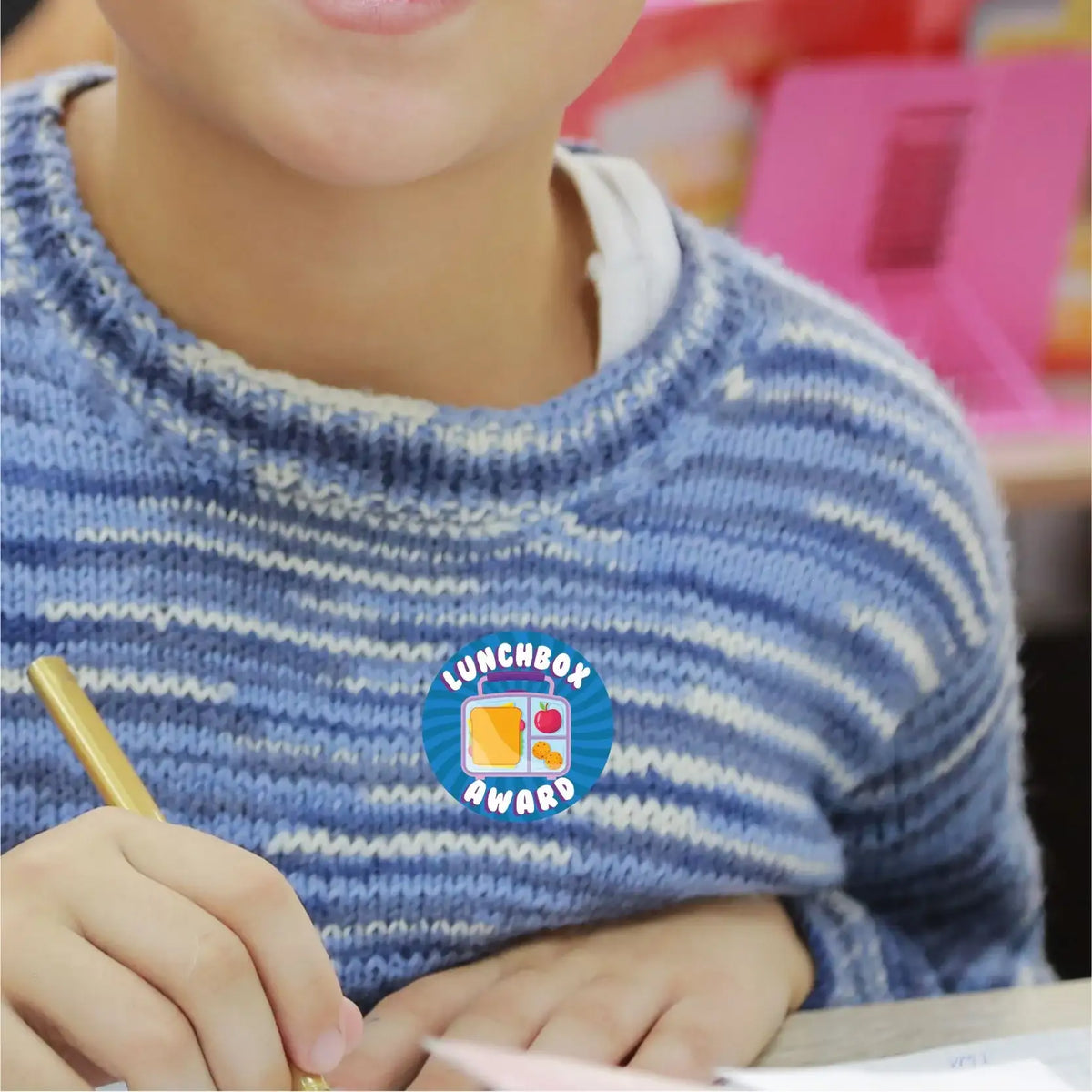 Child wearing Lunchbox Award reward sticker in classroom, holding pencil, striped sweater