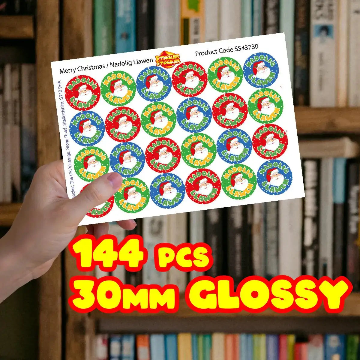 Hand holding Christmas Santa reward stickers sheet, 144 pieces, 30mm glossy, in front of books.